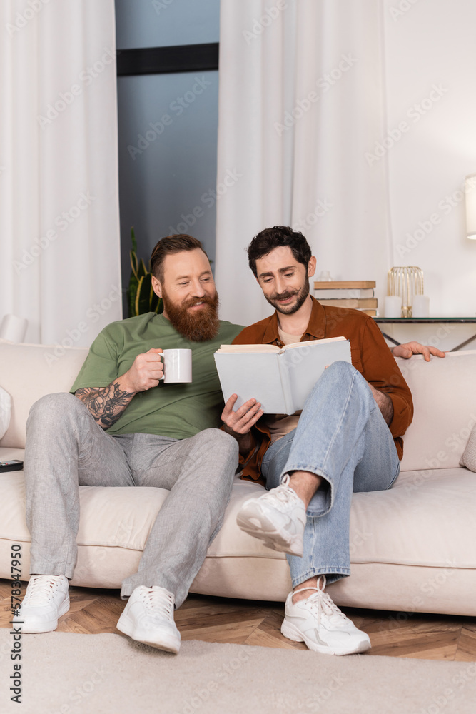 Positive gay couple with coffee reading book at home.