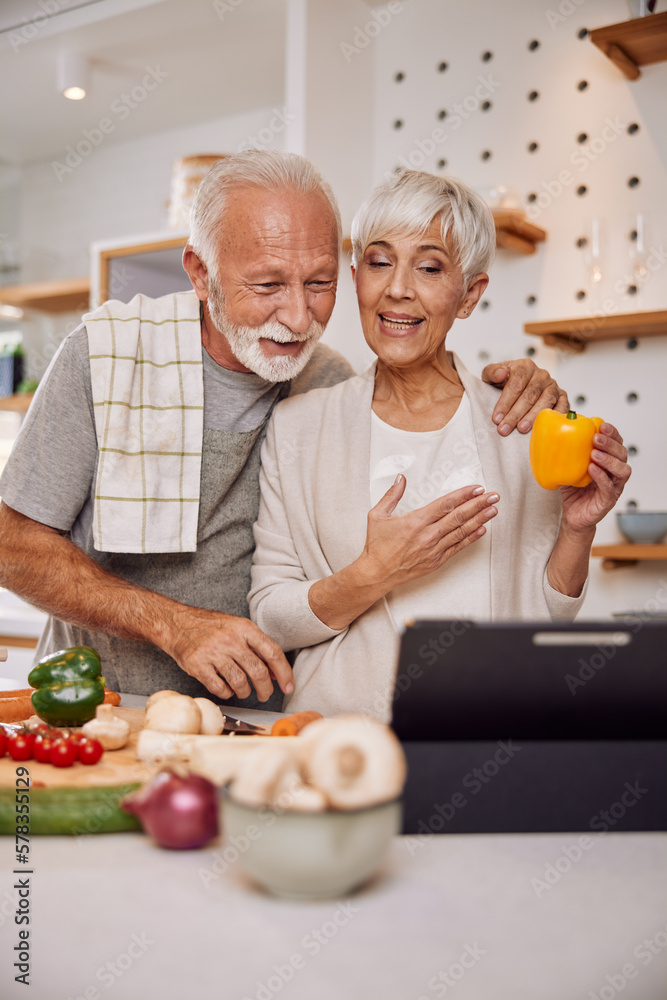 Smiling elderly vegan couple preparing a fresh vegetable salad in the kitchen, using a tablet Smiling elderly vegan couple preparing a fresh vegetable salad in the kitchen, using a tablet