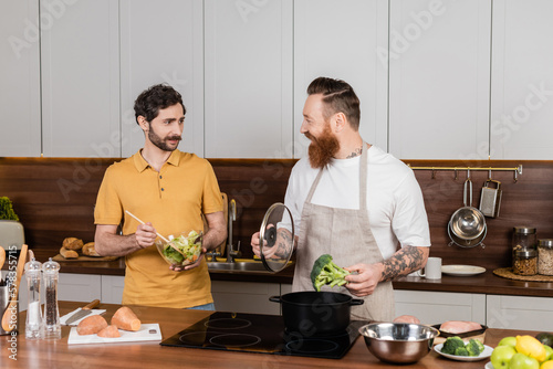 Homosexual man holding salad near partner cooking broccoli in kitchen.
