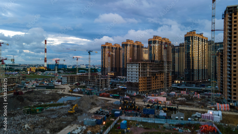 Aerial view of a construct residential high-rise buildings, cranes, steel, concrete, cargo, work next to park and highway in the St. Petersburg at rainy summer evening, shadows