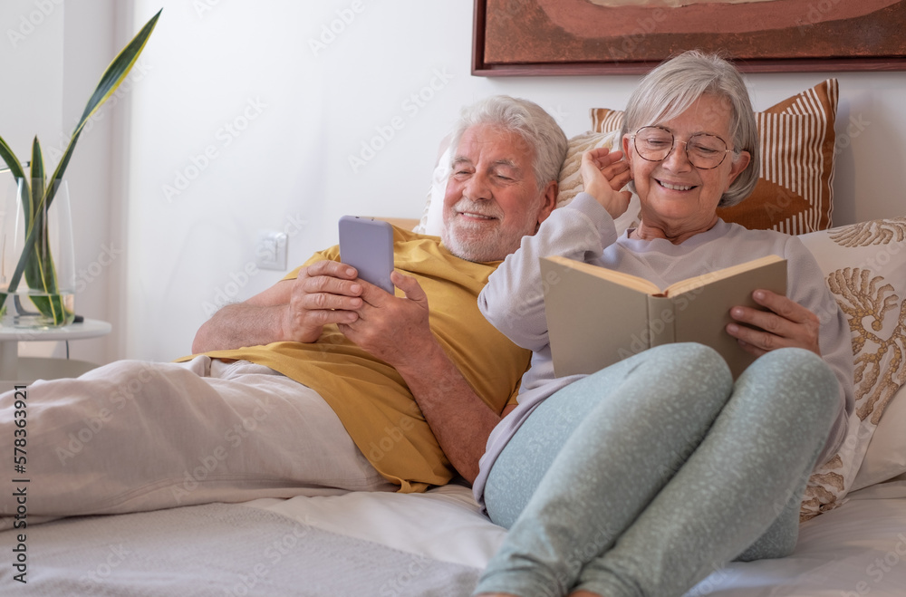 Portrait of happy senior couple together in bed, woman reading book, man using phone Portrait of happy senior couple together in bed, woman reading book, man using phone
