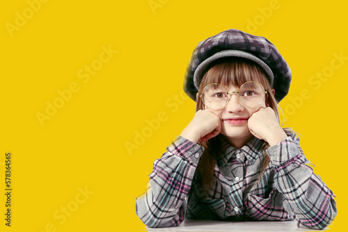 A ten-year-old girl in a plaid hat and a plaid shirt props up her chin with her hand, wearing glasses, close-up, yellow background © MARYIA