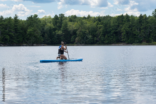 Young fit Caucasian woman in swimming suit and life vest paddle boarding on lake in Northern Ontario. Active lifestyle, adventure, travel, camping, fitness, exercise, water and summer sports concept.