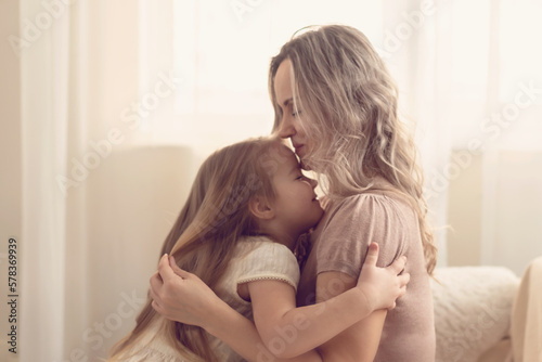 Smiling preschool girl sitting on mommies lap at home. Carefree mom and little girl laughing while playing at home. Love, warm family relationships. Happy kid with mum spending free time together