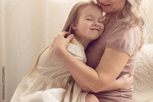 Smiling preschool girl sitting on mommies lap at home. Carefree mom and little girl laughing while playing at home. Love, warm family relationships. Happy kid with mum spending free time together