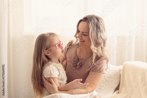 Smiling preschool girl sitting on mommies lap at home. Carefree mom and little girl laughing while playing at home. Love, warm family relationships. Happy kid with mum spending free time together