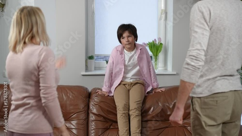 Disobedient boy sitting on couch as unrecognizable parents gesturing scolding kid indoors. Portrait of misbehaving Caucasian son with angry mother and father at home. Individuality and lifestyle