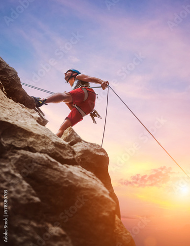 Muscular climber man in protective helmet abseiling from cliff rock wall using rope Belay device and climbing harness on evening sunset sky background. Active extreme sports time spending concept.