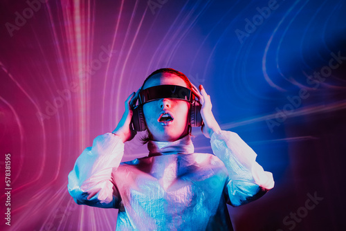 Portrait of sensitive dancing woman in futuristic sunglasses and headphones in pink and blue neon light. Music lover. Silent disco. Woman on the background of music vibes background. Futuristic Party. photo