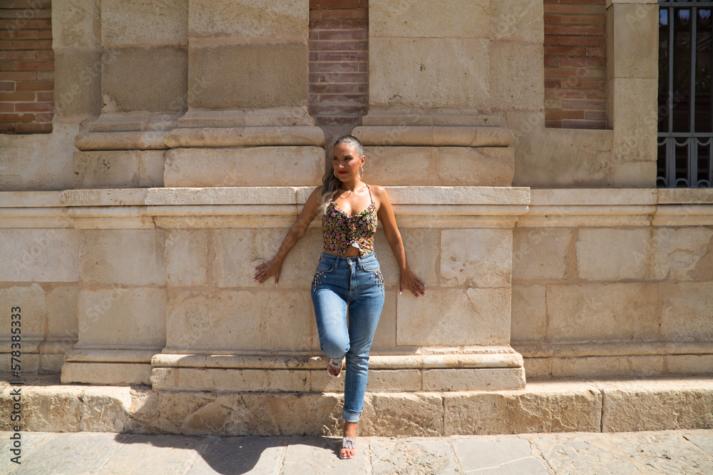 Beautiful young blonde Spanish woman is leaning against the wall of an old building in Seville. The woman is dressed in jeans. The woman is sightseeing and visiting spain.