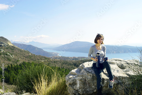 A young woman is about to pour a drink from a thermos into a cup, sitting on a stone against the backdrop of a magnificent view of the mountains, forest and sea. Portrait of a tourist on a halt