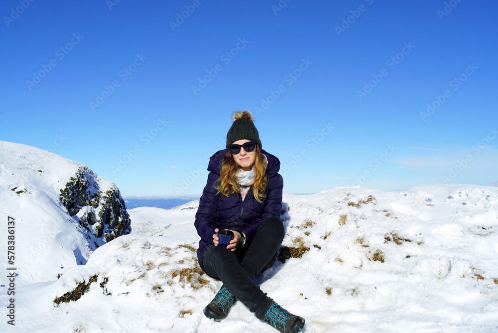 A young woman sits on top of a snow-covered mountain with a cup of drink in her hands. Rest in the ski resort of Montenegro.