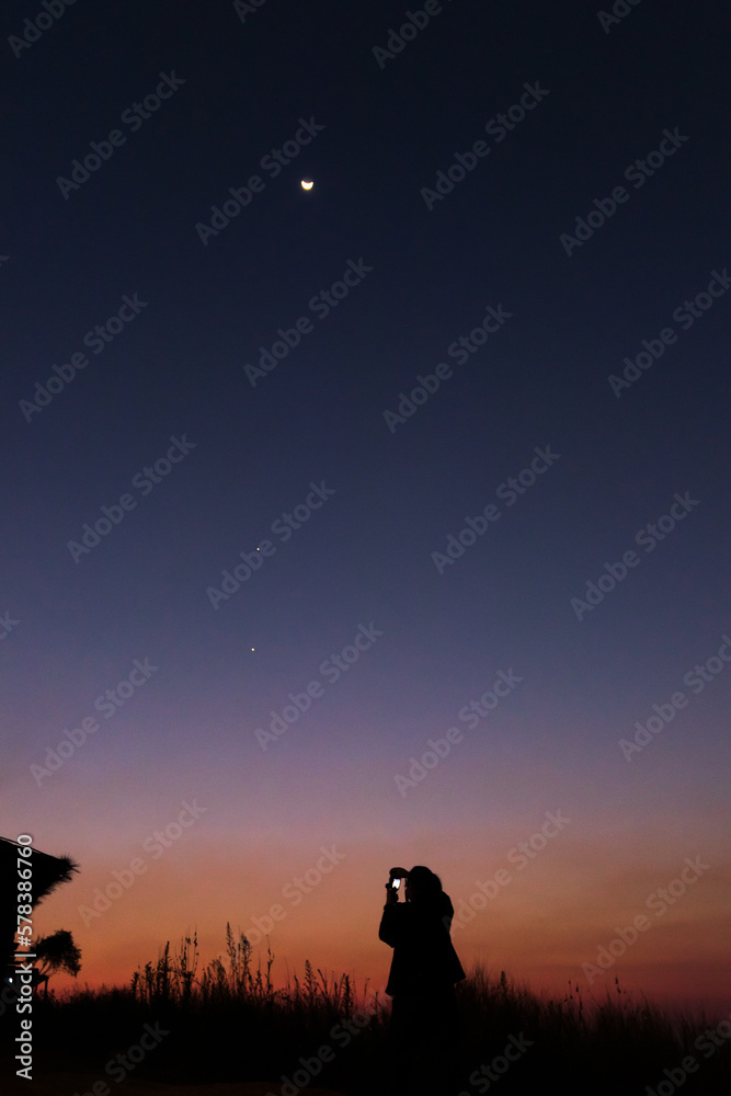 Women taking picture of nature observing evening sky with stars, planets and crescent moon. Moon meets Jupiter and Venus in the twilight sky.