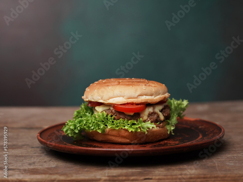 Tasty Burger lies on clay plate against wooden table. A Juicy green Salad leaf and a red Tomato