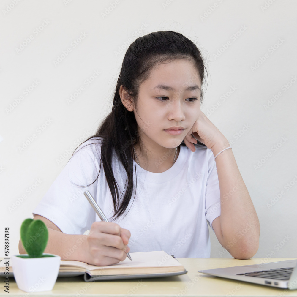 Asian girl is studying on a laptop and writes in a notebook with intention,Online learning, stay home, New normal, Covid-19 coronavirus, Social distancing, Internet learning.. Asian girl is studying on a laptop and writes in a notebook with intention,Online learning, stay home, New normal, Covid-19 coronavirus, Social distancing, Internet learning..
