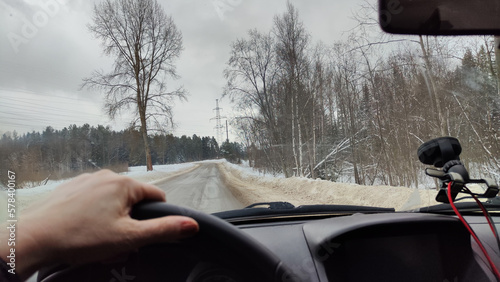 Hand of woman on the steering wheel in a car and forest full of snow on the side of ice road on a winter day. Woman driving a car in winter travel in landscape with snow covered woods