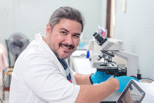 A cheerful male laboratory technician making a thumbs up while checking a swab sample with a microscope. Working at a research facility of hospital lab. Wearing a bouffant cap and face mask.
