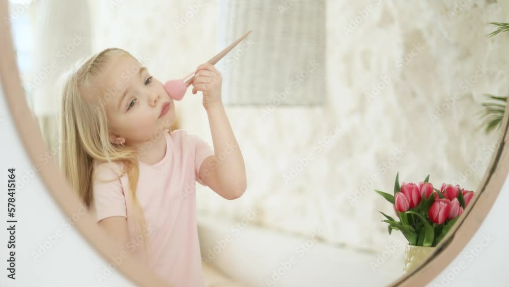 Cute little girl playing like doing daily make up in the morning holding brush putting face powder for chicks and looking at her mirror reflection in the light bedroom Happy childhood concept Cute little girl playing like doing daily make up in the morning holding brush putting face powder for chicks and looking at her mirror reflection in the light bedroom Happy childhood concept