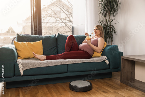 Pregnant Woman Relaxing On Sofa While Robotic Vacuum Cleaner Cleaning At Home