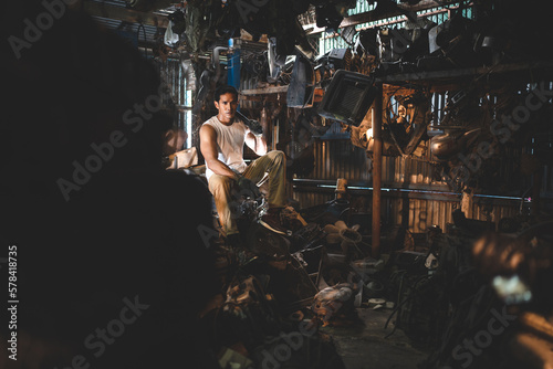 Young and handsome muscular and strop scrap material man in sleeveless shirt sitting on material in junkyard while looking away while taking rest and break from work during in warehouse