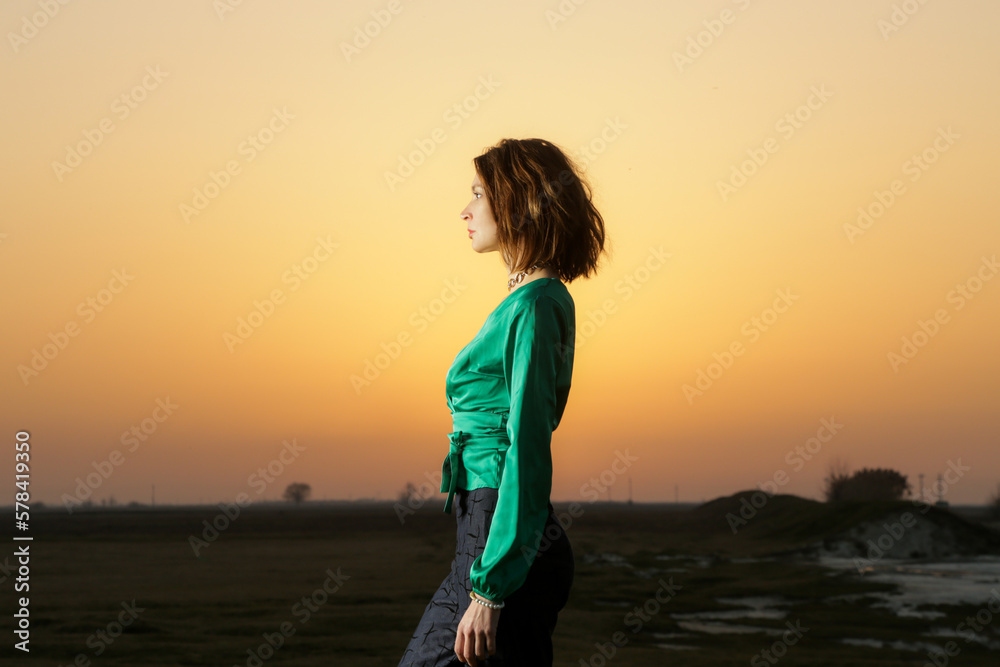 Fashion style portrait of young stunning woman posing in green silk blouse and denim trousers during sunset time 