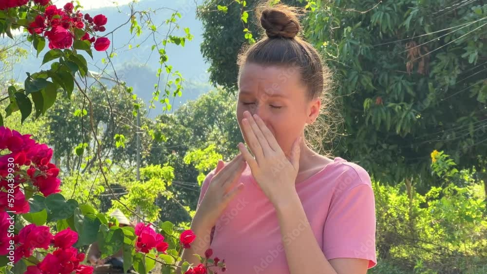 Portrait of beautiful young allergic woman is suffering from pollen allergy or cold on natural flower flowering tree background at spring or sunny summer day sneezes, blowing her runny nose rubs eyes