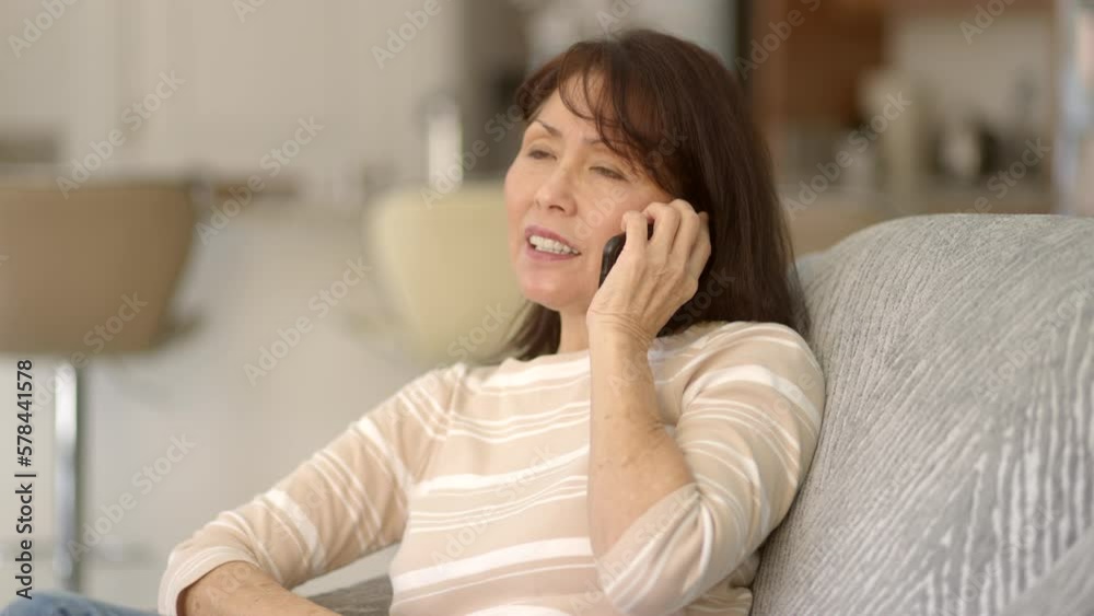 Smiling attractive stylish woman enjoying talking on mobile cell phone smiling brushing hair from face laughing, sitting on sofa, kitchen background. Asian female using internet communication. Smiling attractive stylish woman enjoying talking on mobile cell phone smiling brushing hair from face laughing, sitting on sofa, kitchen background. Asian female using internet communication.