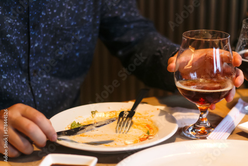 After meal concept. Dirty dishes from the tables in restaurant. Empty dirty plate after meal with fork inside next to napkins with chopsticks, male hand. Man holding glass with beer or whiskey