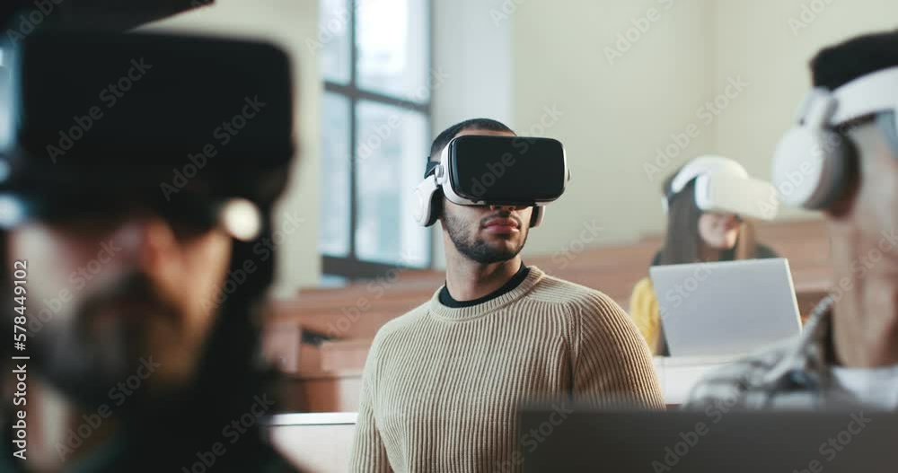 Multiethnic males and female students in VR glasses listening to lection at University and turning heads left and right. Hi-tech education. Mixed-races young people learning with virtual reality.