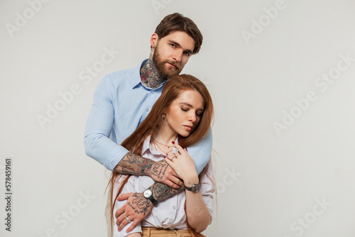 Fashion beautiful lovers couple in the studio. Pretty redhead woman and handsome hipster man with tattoos hug on a white background