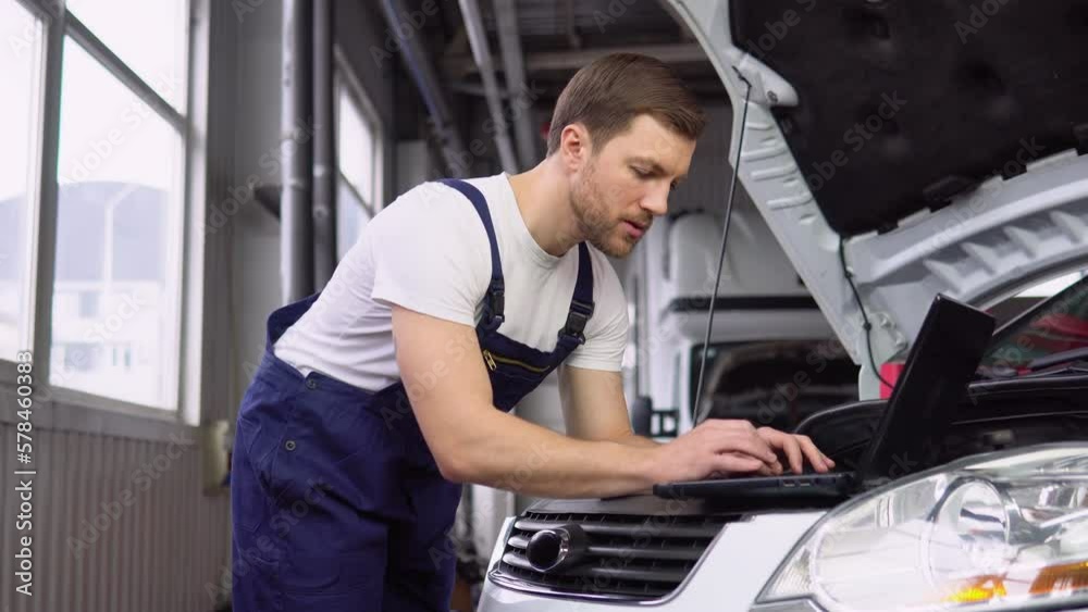 Professional car mechanic working in auto repair service with laptop. Adult man in uniform works in the automobile salon Professional car mechanic working in auto repair service with laptop. Adult man in uniform works in the automobile salon