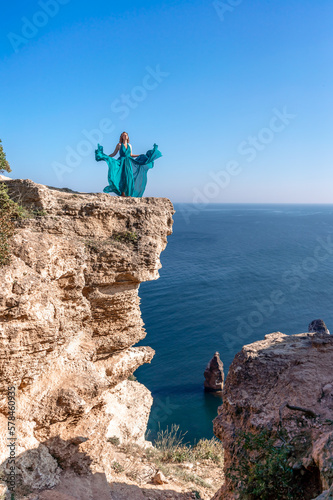 A girl with loose hair in a long mint dress descends the stairs between the yellow rocks overlooking the sea. A rock can be seen in the sea. Sunny path on the sea from the rising sun