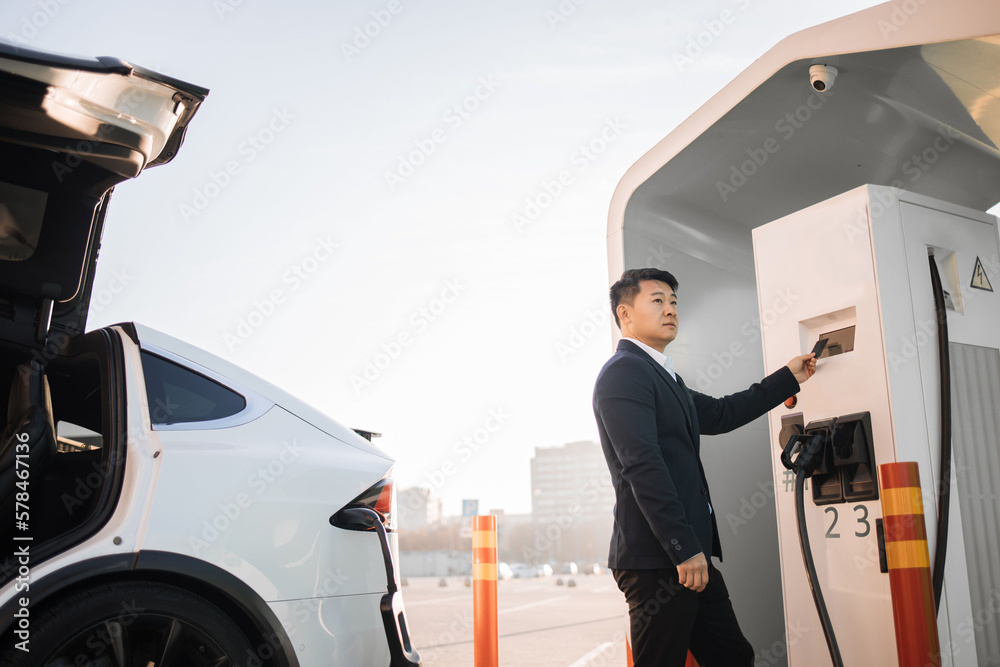 Side view of successful asian businessman paying with credit card for energy at charging station. Stylish man standing outdoors near luxury electro car and using cashless payment. Side view of successful asian businessman paying with credit card for energy at charging station. Stylish man standing outdoors near luxury electro car and using cashless payment.