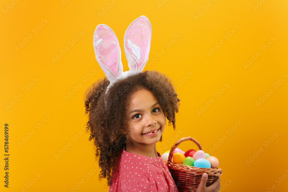 A Black girl with rabbit ears on her head with a basket full of colored eggs. A Black girl with rabbit ears on her head with a basket full of colored eggs.