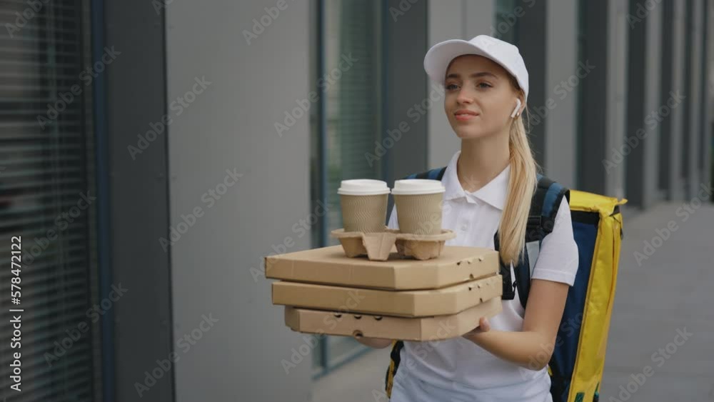 Caucasian Positive Woman Courier Wearing White Uniform, Carrying the Backpack, Holding a Pizza and Coffee in Her Hands for a Client, Wearing Headphones. Food Delivery. People and Professions Caucasian Positive Woman Courier Wearing White Uniform, Carrying the Backpack, Holding a Pizza and Coffee in Her Hands for a Client, Wearing Headphones. Food Delivery. People and Professions
