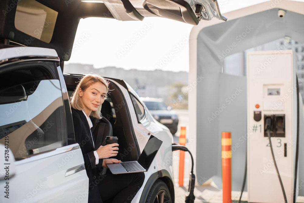 Attractive woman with blond hair enjoying coffee-to-go and working on portable laptop while charging electric car at outdoors station. Concept of business people, technology and ecological transport.