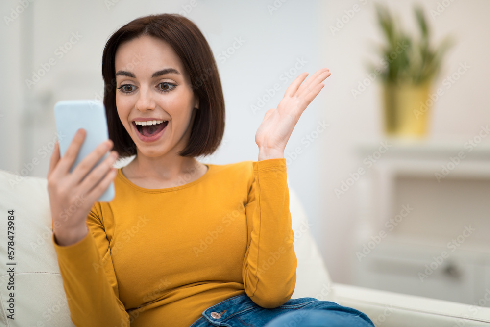 Emotional young woman sitting on couch at home, using smartphone Emotional young woman sitting on couch at home, using smartphone
