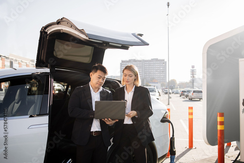 Two focused multiracial business partners in formal attire working on wireless laptop while waiting for luxury electric car to be charged. Concept of people, ecology and technology.