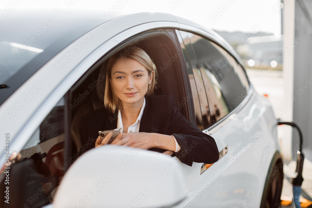 Attractive caucasian woman with modern smartphone in hands sitting inside luxury car while recharging battery on auto station. Business lady in stylish suit working online during trip break. Attractive caucasian woman with modern smartphone in hands sitting inside luxury car while recharging battery on auto station. Business lady in stylish suit working online during trip break.