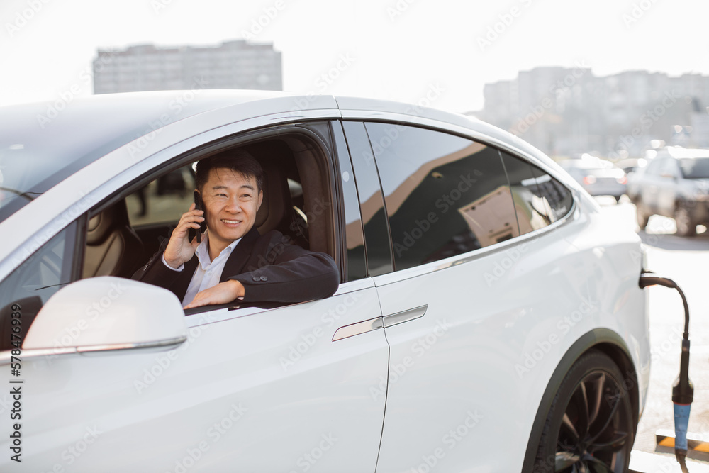 Smiling office worker in business suit chatting on smartphone while his electric car being charged through power cable supply at station. Happy asian man enjoying talks while refilling EV with energy.