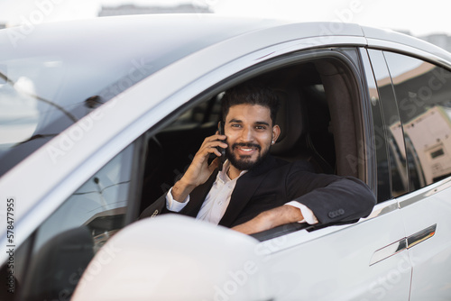 Attractive bearded man in formal suit talking on smartphone while sitting inside brand new auto. Well-dressed indian man smiling happily while looking out of open car window outdoors.