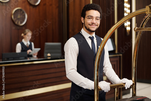 Young elegant bellboy in uniform pushing cart with clients baggage while moving along hotel lounge and looking at camera photo