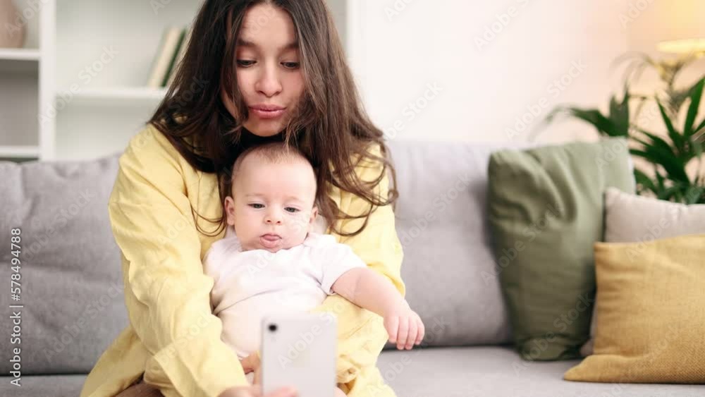 Portrait of cute young woman mother hug her little baby and grimacing taking selfie photograph saving great memories on smartphone mobile phone at light home Happy motherhood concept Portrait of cute young woman mother hug her little baby and grimacing taking selfie photograph saving great memories on smartphone mobile phone at light home Happy motherhood concept