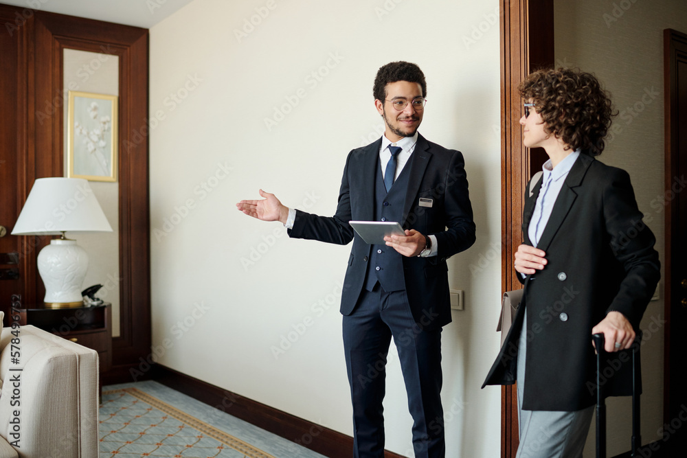 Fototapeta premium Young confident head manager of luxurious hotel showing room to female guest in smart casualwear with suitcase listening to him