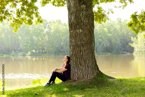 Teen young girl woman sitting on grass under huge high big old oak,trunk tree near lake,river, meditating,relaxing and smiling in National Park.inspiration nature beauty harmony landscape outdoors