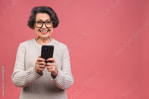 Happy mature senior woman holding smartphone using mobile online apps, smiling old middle aged grandmother texting sms message chatting on phone isolated over pink background.