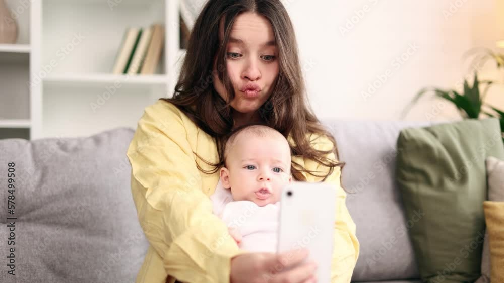 Portrait of cute young woman mother hug her little baby and grimacing taking selfie photograph saving great memories on smartphone mobile phone at light home Happy motherhood concept Portrait of cute young woman mother hug her little baby and grimacing taking selfie photograph saving great memories on smartphone mobile phone at light home Happy motherhood concept