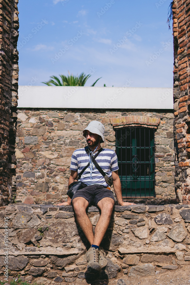 Young Latin man with a bucket hat sitting on colonial ruins thinking about what might have been. Vertical photo.
