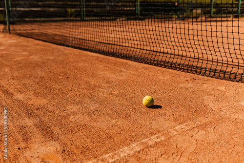 Light orange empty outdoor clay court surface dry grungy ground baseline detail for playing tennis with net in sunny day with yellow green ball, gravel texture background, copy space for text   © Volodymyr