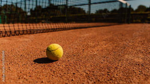 Light orange empty outdoor clay court surface dry grungy ground baseline detail for playing tennis with net in sunny day with yellow green ball, gravel texture background, copy space for text    © Volodymyr